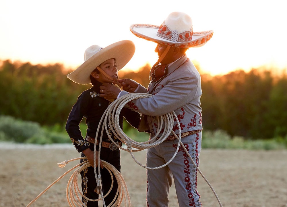 Un hombre y un niño vestidos con el traje tradicional Charro, sosteniendo cuerdas y parados uno al lado del otro.